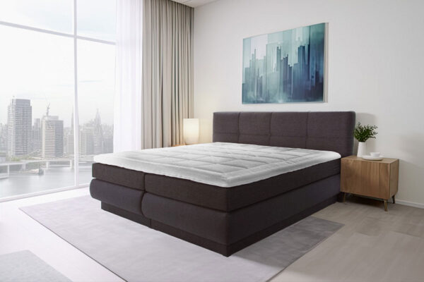 Modern boxspring bed with dark frame and light mattress topper in a bright bedroom with city view.