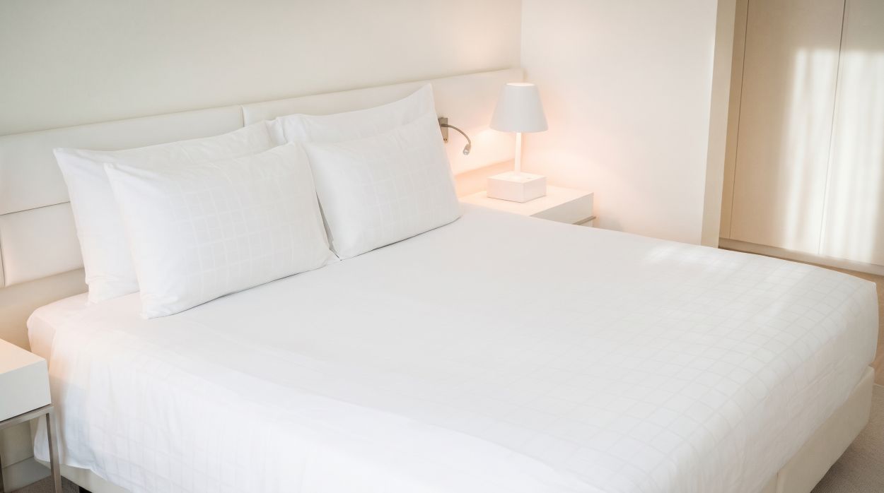 Een strak opgemaakt tweepersoonsbed met wit bedtextiel en kussens in een lichte hotelkamer setting, passend bij brandvertragende dekbedden.